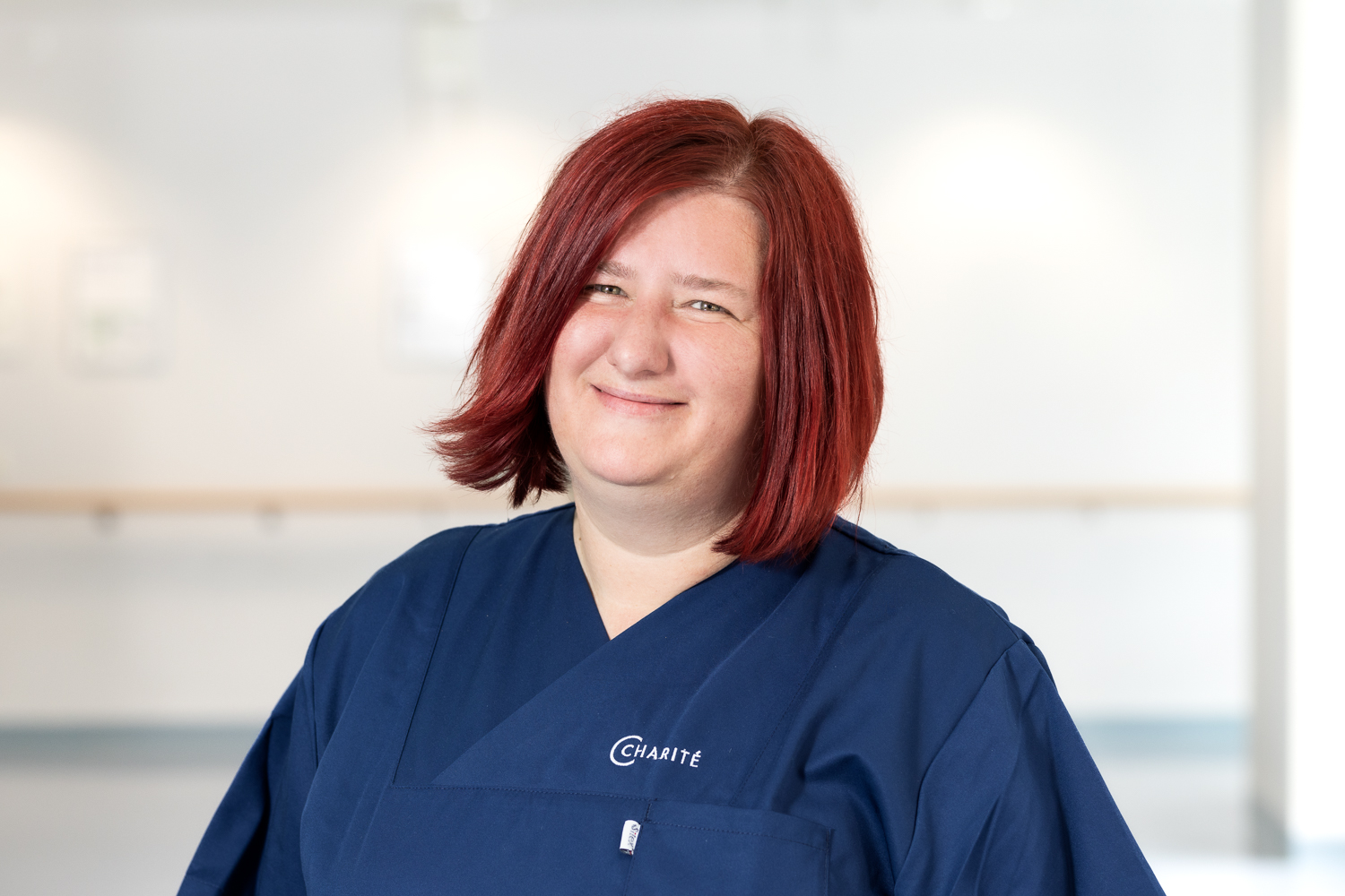 Eine lächelnde Frau mit rotem Haar trägt eine dunkelblaue Berufskleidung. Sie steht in einem hellen Raum mit unscharfen Details im Hintergrund, die auf eine gesundheitsbezogene Umgebung hinweisen.