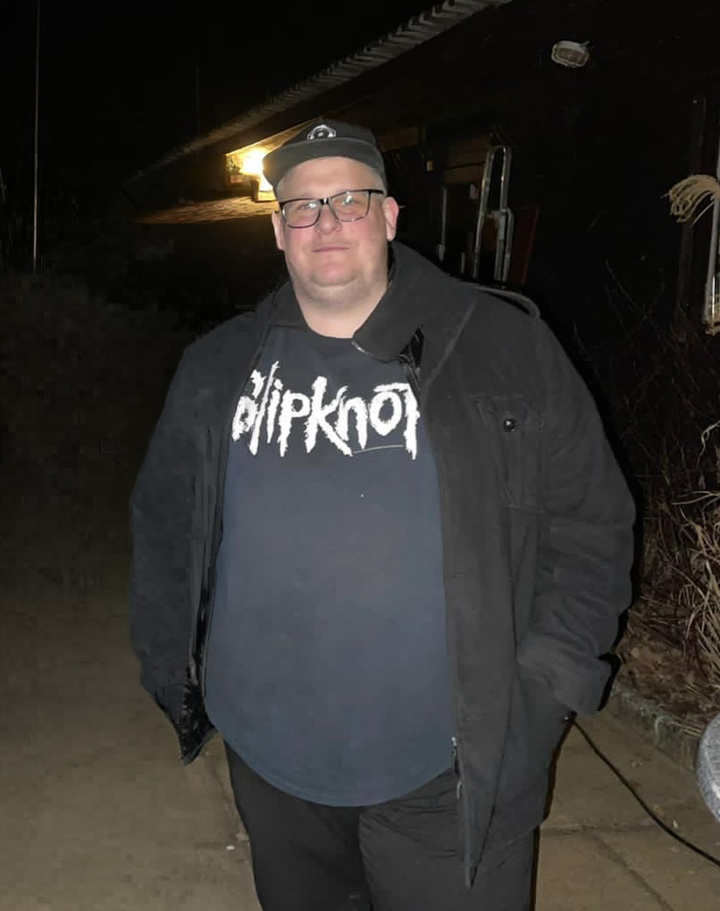 Ein Mann steht in der Nacht draußen. Er trägt ein schwarzes Shirt mit dem Schriftzug „Slipknot“ und eine schwarze Jacke. Im Hintergrund ist ein Gebäude sichtbar, das schwach beleuchtet ist. Die Umgebung wirkt dunkel und ruhig.
