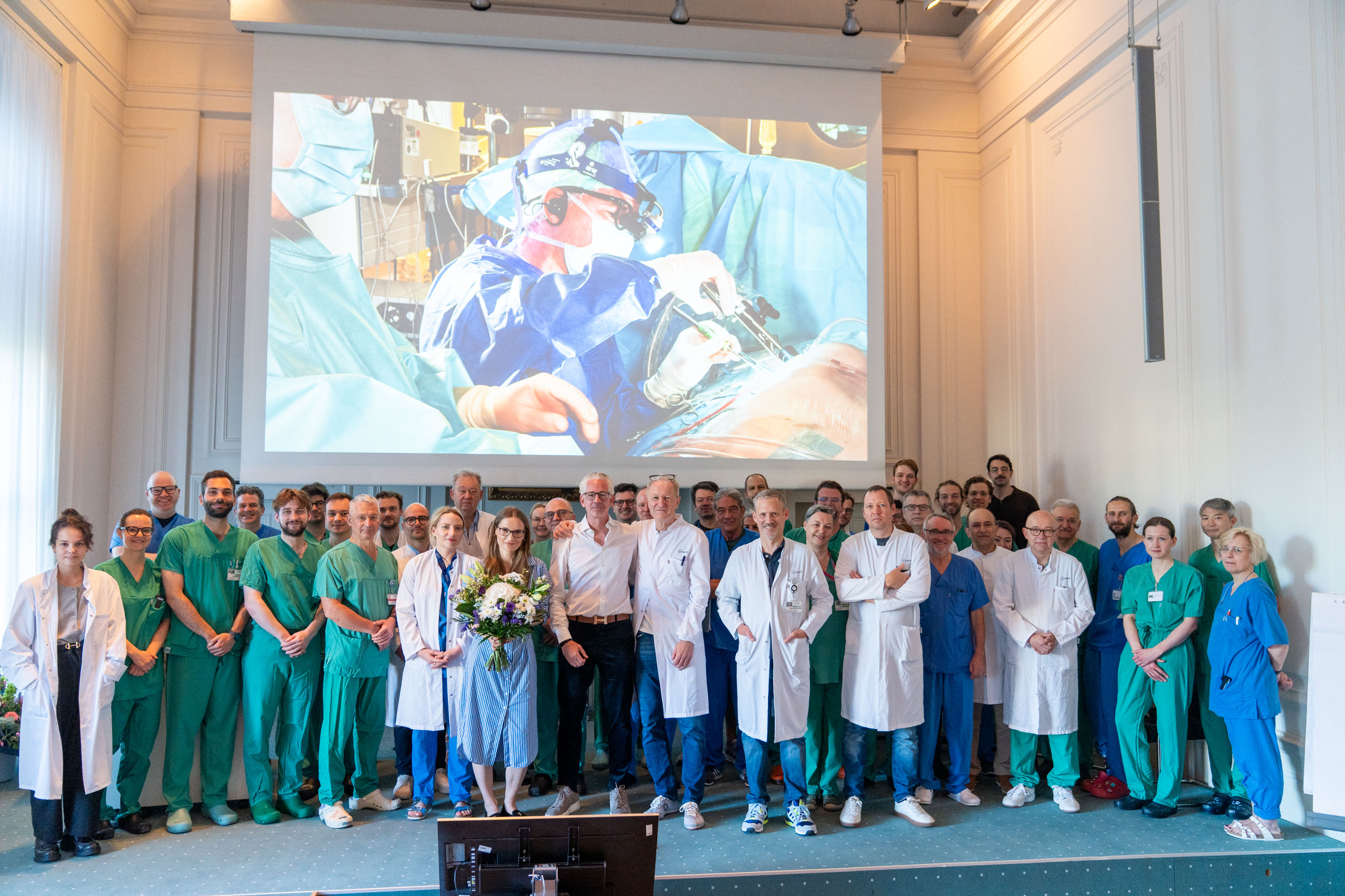 Eine Gruppe von Medizinern und Mitarbeitern steht zusammen in einem Saal. Im Hintergrund ist ein Bild einer chirurgischen Operation auf einer Leinwand zu sehen. Die Personen tragen unterschiedliche medizinische Kleidung und lächeln in die Kamera, während sie eine Blumenstrauß präsentieren.