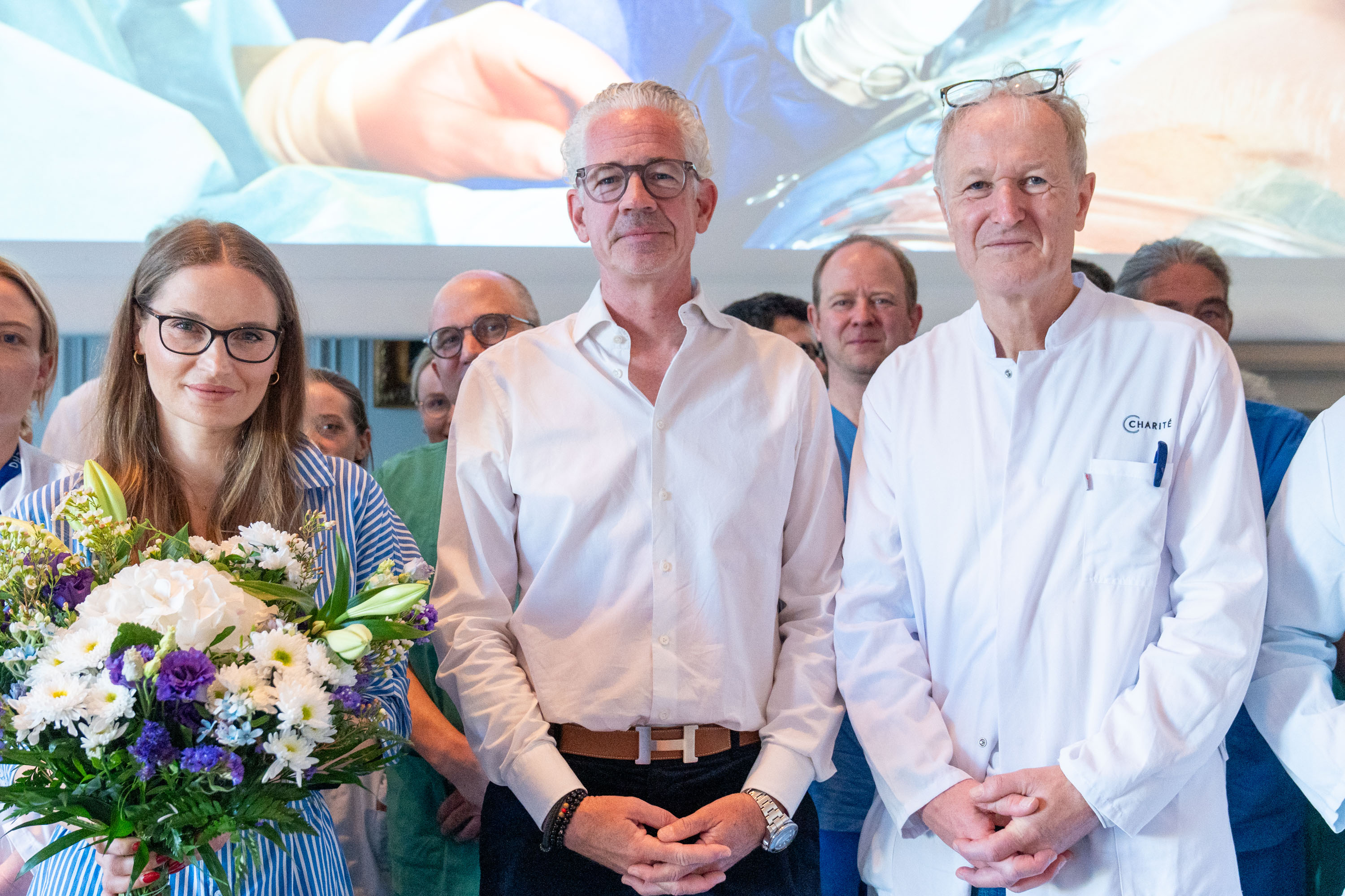 Eine Gruppe von medizinischem Personal steht zusammen, darunter eine Frau mit Blumenstrauß, ein Mann in Hemd und Kragen, und ein Arzt in weißen Kittel. Im Hintergrund ist eine Präsentation mit einer medizinischen Szene sichtbar. Alle wirken glücklich und stolz.