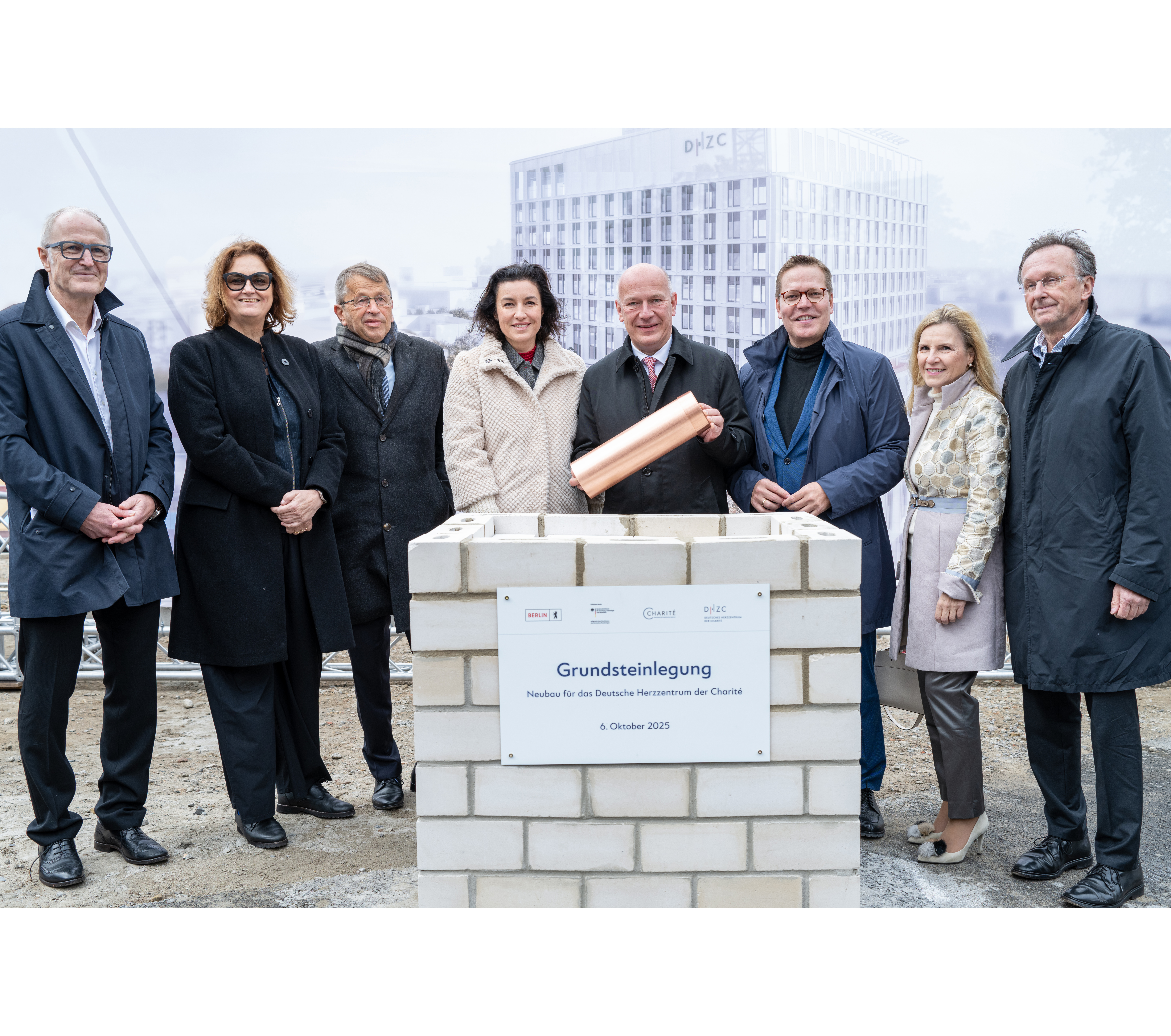 Eine Gruppe von sieben Personen steht um einen Grundstein, der mit einer Plakette versehen ist. Der Grundstein trägt die Aufschrift „Grundsteinlegung Neubau für das Deutsche Herzzentrum der Charité, 6. Oktober 2025“. Im Hintergrund ist eine bauprojekte Darstellung sichtbar.