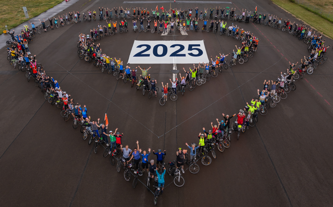 Eine Gruppe von Radfahrern bildet in einer herzförmigen Anordnung die Zahl "2025" auf einem Asphaltplatz. Die Teilnehmer tragen verschiedene bunte Radkleidung und heben die Arme zur Feier. Die Szene vermittelt Gemeinschaft und Vorfreude auf das Jahr 2025.